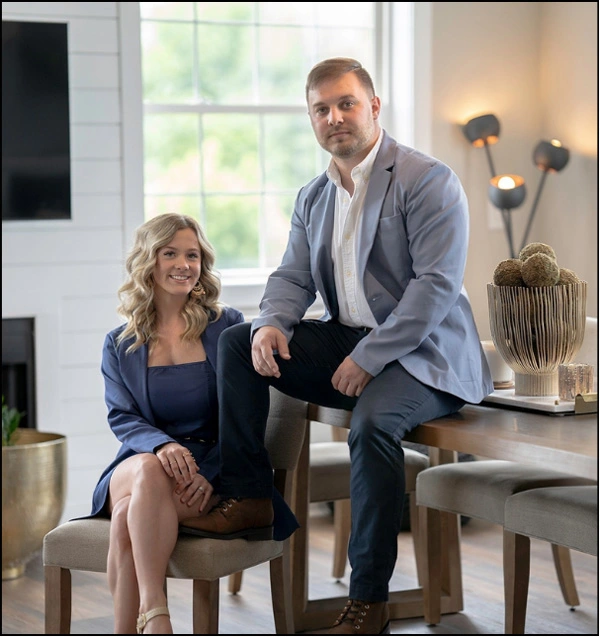 A professional portrait of a man and a woman, both wearing blue blazers, posing in a well-lit, modern dining and living room area, suitable for a real estate or business team.