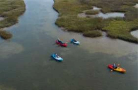Drone shot of four brightly colored kayaks (red, blue, and yellow) paddling down a shallow, winding river channel surrounded by dense, brown and green marsh grass and wetlands.