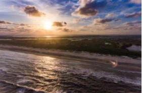 Aerial view of a wide, wet beach and churning ocean waves at sunset, with the bright sun reflecting intensely off the water and illuminating the dark coastal marshland stretching toward the horizon under a cloudy sky.