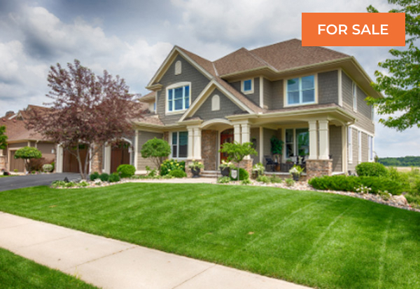 Exterior view of a large, two-story luxury suburban home painted in shades of tan and brown, featuring a covered front porch with stone columns, a spacious, bright green manicured lawn, and a sidewalk leading to the street, with a "FOR SALE" tag.