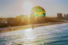 Aerial view of a person parasailing with a brightly colored yellow, blue, and green parachute over a wide beach and blue ocean waves, with a coastal city skyline and bright, lens-flared sun visible in the background.