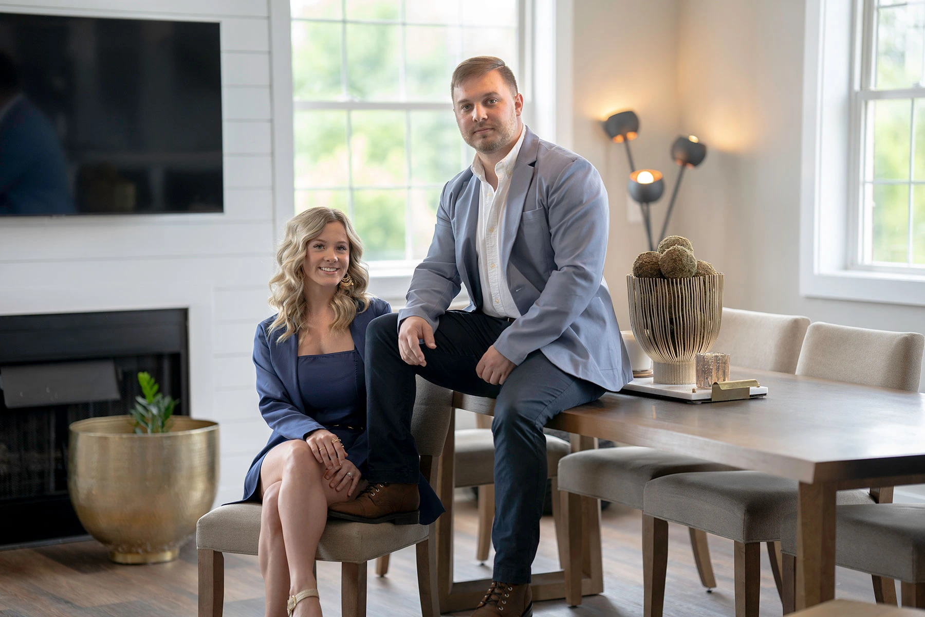 Professional portrait of a man and a woman, both wearing blue blazers, sitting and posing near a dining table in a brightly lit, modern interior living space with white walls.