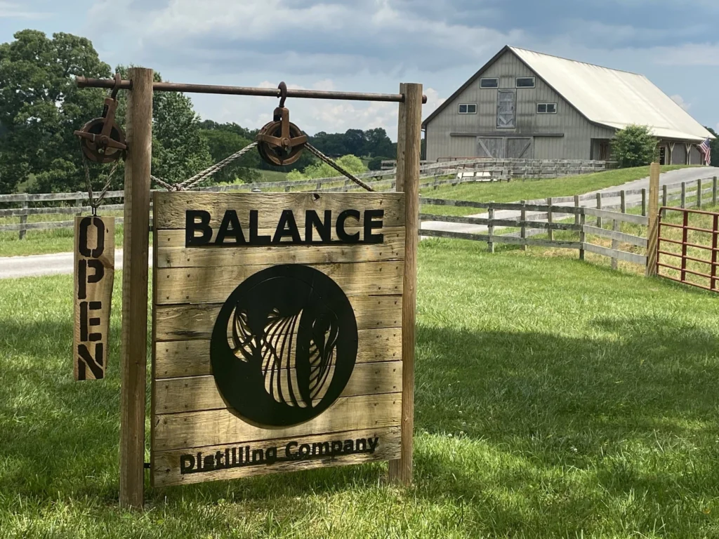 Rustic entrance sign at Balance Distilling Company in a grassy field. In the background is a large light gray wooden barn where the distillery is located.