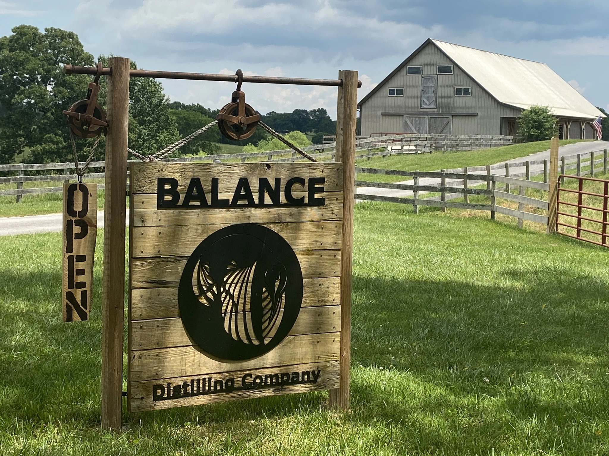 Rustic entrance sign at Balance Distilling Company in a grassy field. In the background is a large light gray wooden barn where the distillery is located.