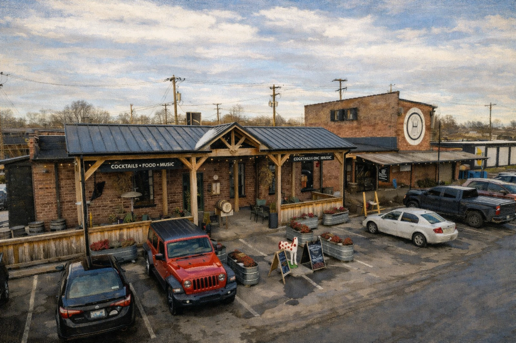 Exterior view of Barrel House Distilling Co with covered patio seating, parked cars, and brick buildings under a partly cloudy sky.