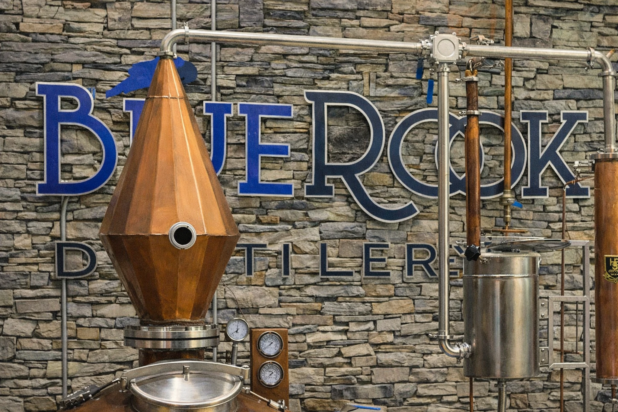 Copper still and distillation equipment inside Blue Rook Distillery, viewed through a large interior window with stone walls and seating area.