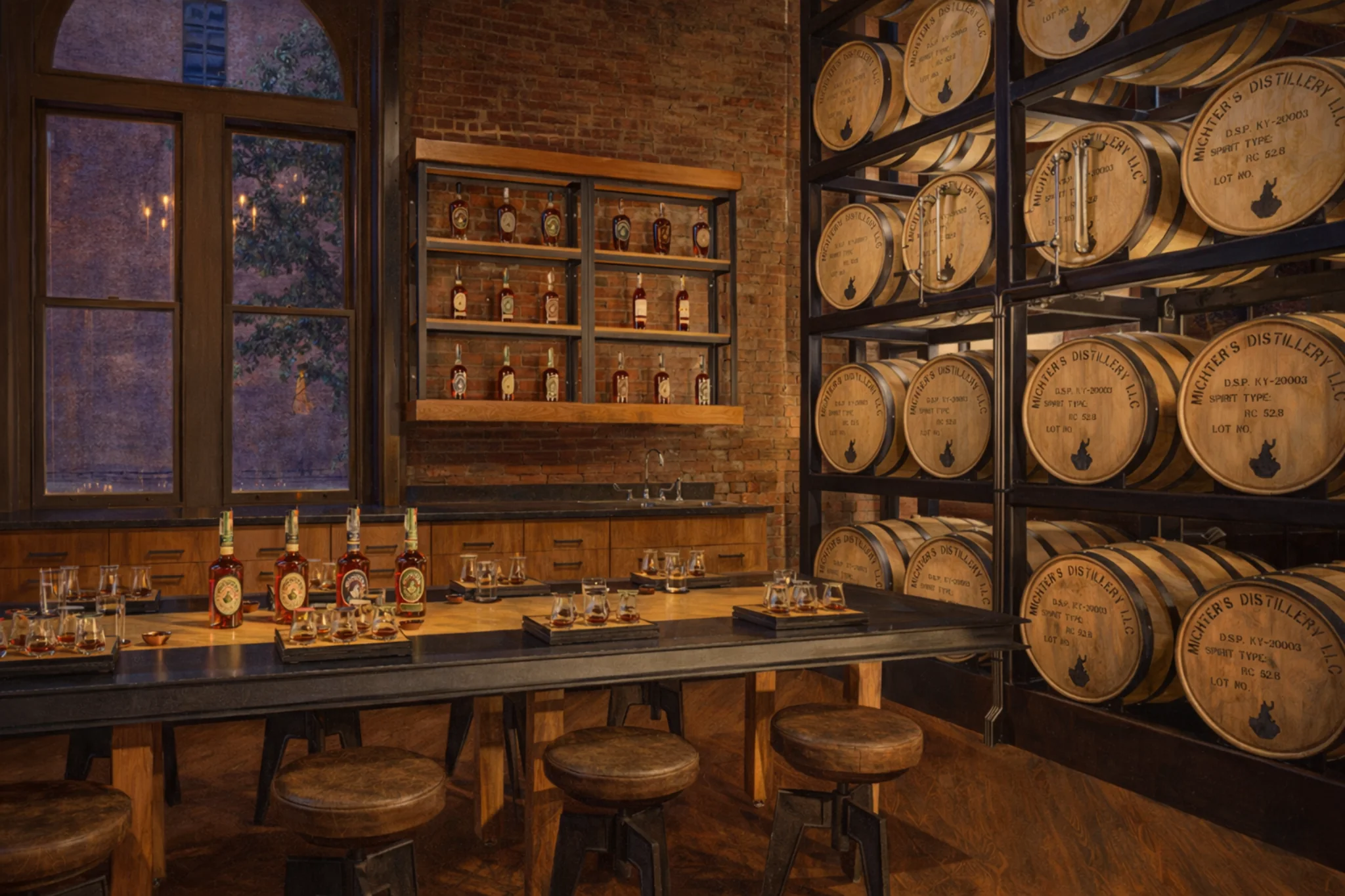 Michter’s Fort Nelson Distillery tasting room in Louisville, Kentucky with a long tasting table set with bourbon glasses and a wall of Michter’s barrels.