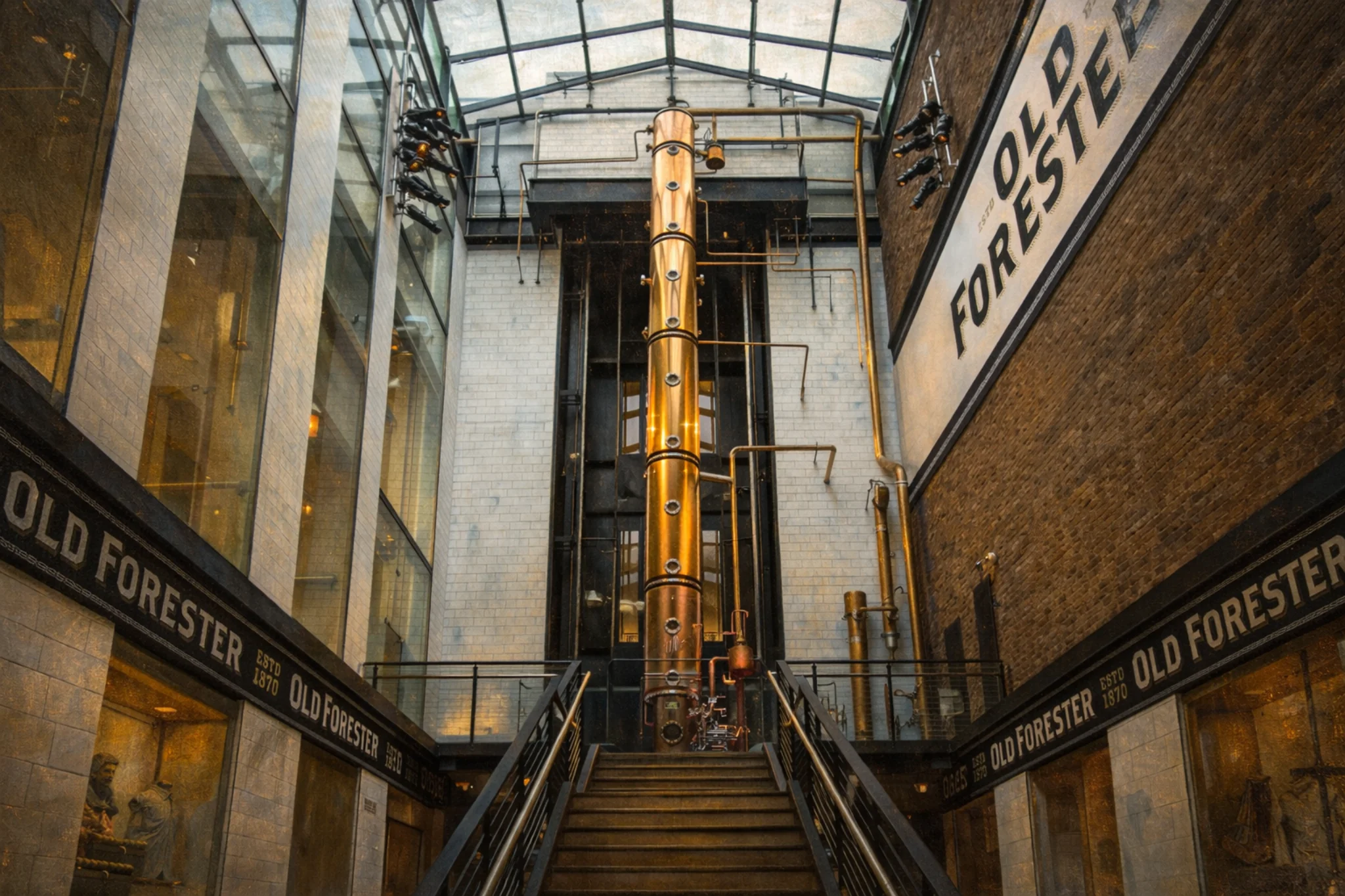Atrium stairway view inside Old Forester Distilling Co. featuring a tall copper column still beneath a glass roof and Old Forester wall signage.
