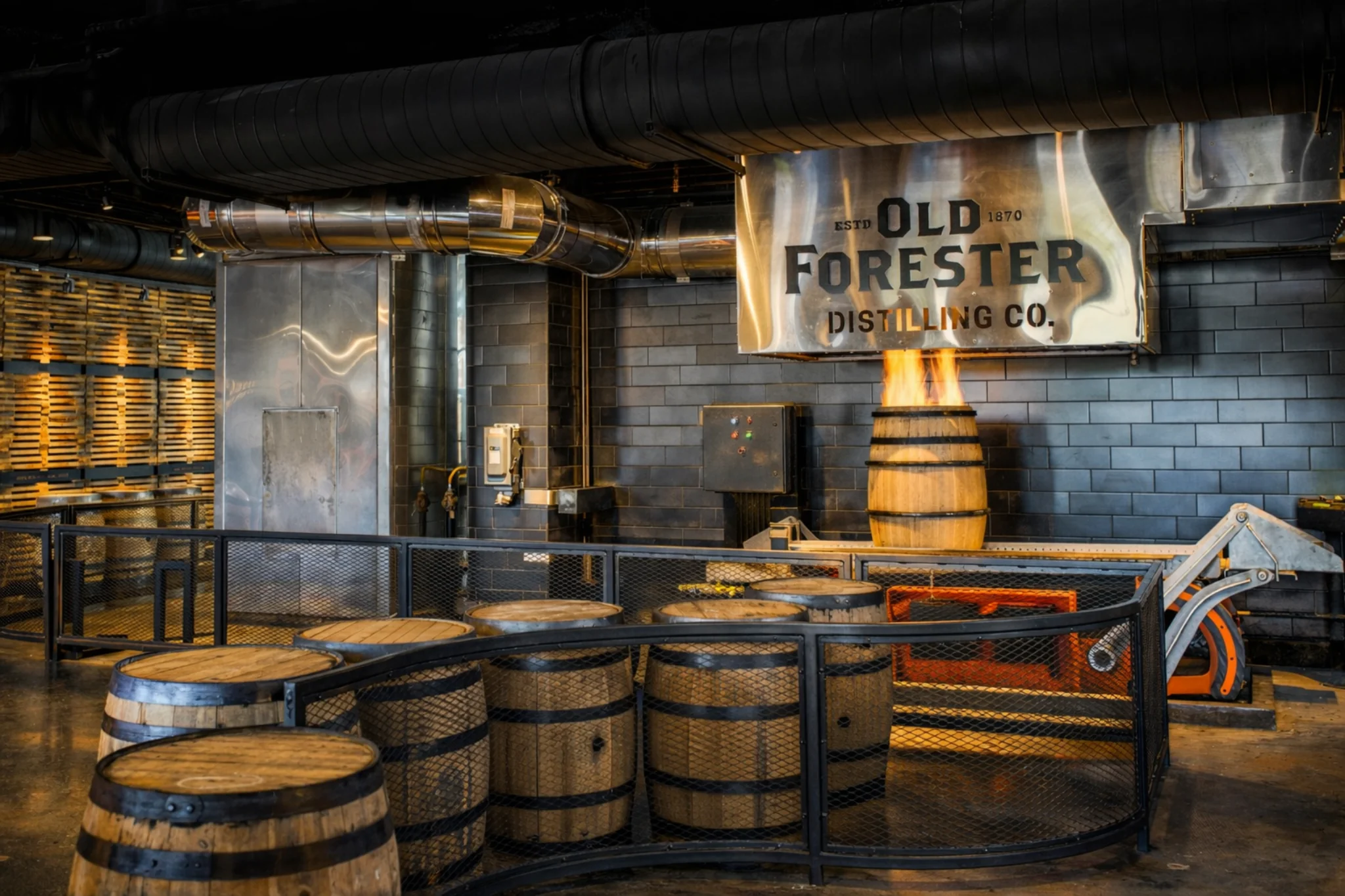 Old Forester Distilling Co. barrel charring display with a flame rising from a barrel beneath an Old Forester sign in an industrial setting.