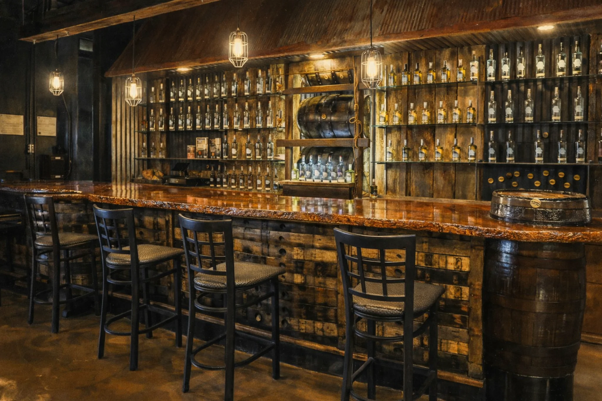 Rustic tasting room bar at Prohibition Craft Spirits with a long wood countertop, barrel-front bar design, and shelves of spirits bottles under warm pendant lighting.