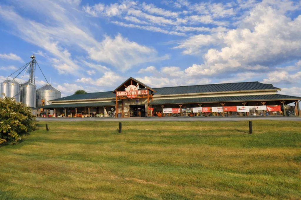 Exterior view of Bardstown Mills with the main storefront, grassy foreground, and silos under a bright sky.